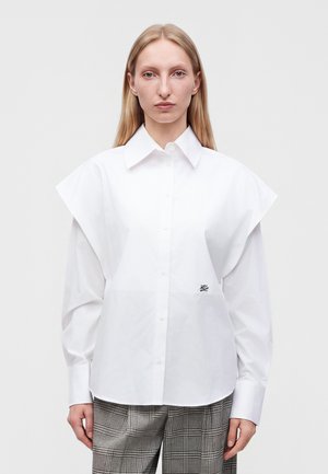 Femme aux longs cheveux blonds portant une chemise blanche boutonnée avec des épaules larges et un pantalon à carreaux gris, debout devant un fond uni.