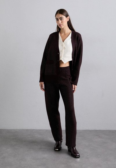 Bordeaux cardigan met stippen, gecombineerd met een crèmekleurige cropped blouse en bijpassende broek. Glanzende zwarte laarzen maken de outfit compleet.