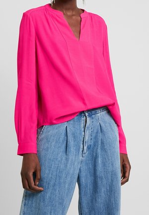 Blouse rose vif à manches longues avec un col en V, coupe ample et texture douce, associée à un pantalon en jean plissé bleu clair.
