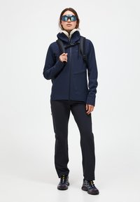 Giacca softshell blu navy con zip frontale, cappuccio e due tasche, abbinata a pantaloni neri e scarpe da trekking multicolori.