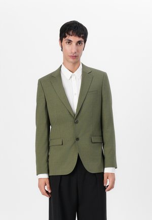 Olivenfarget blazer med teksturert stoff, to frontknapper, spisse krager og to klaffelommer, paret med en hvit skjorte.