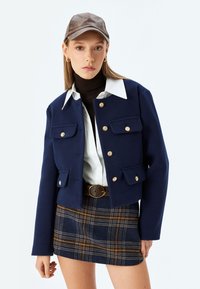 Giacca corta blu marino con bottoni dorati e tasche a pattina, indossata su una camicia bianca e un dolcevita marrone, abbinata a una mini gonna a cuadros.