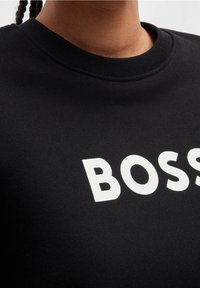 Černá mikina s kulatým výstřihem, která má na přední straně výraznou bílou grafiku "BOSS". Vyrobena z měkkého bavlněného materiálu.