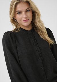 Schwarze Bluse mit langen Ärmeln, Rüschenkragen, Knopfleiste und strukturiertem Stoff. Mit Puffärmeln und dezenter Schimmer.