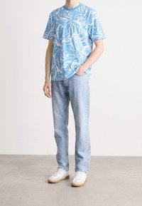 Uomo che indossa una maglietta a righe blu e bianca con motivo marmorizzato, jeans azzurri chiari, sneaker bianche, in piedi con una mano in tasca su uno sfondo neutro.