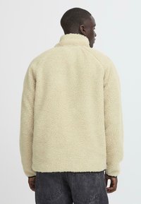 Fleece jacket in cream, featuring a textured surface, high collar, and long sleeves. The hem is slightly curved with elastic cuffs.