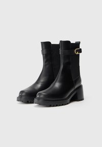 Par de botas de cano curto em pele preta com saltos grossos, painéis laterais elásticos e uma fivela decorativa dourada sobre um fundo claro.