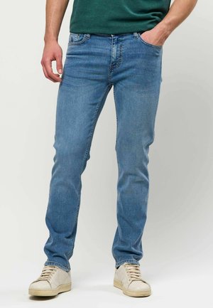Mies, joka pukeutunut vaalean sinisiin slim fit -farkkuihin ja beigeihin lenkkareihin, seisoo käsi yhdessä taskussa yksivärisen taustan edessä.