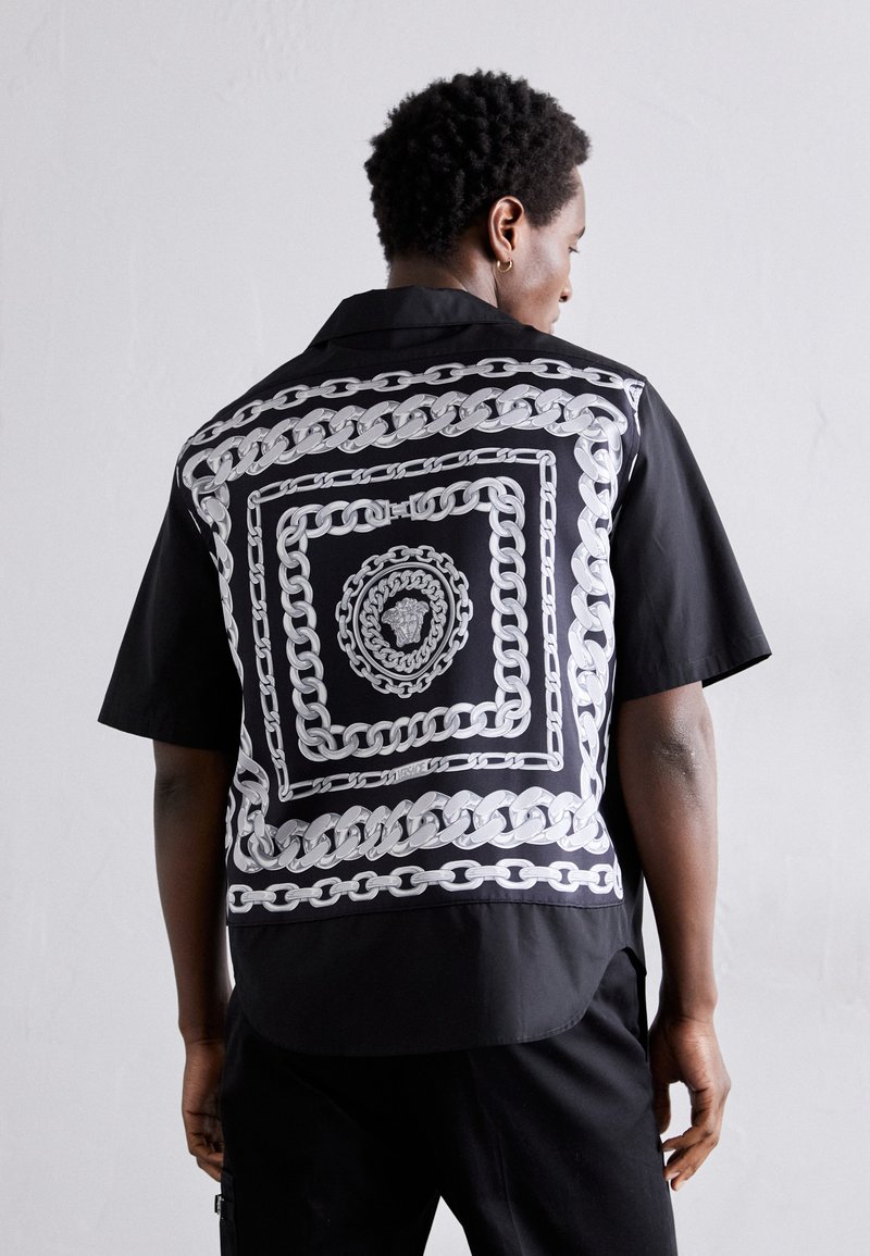 Chemise noire à manches courtes avec un grand motif de chaîne argentée au dos, texture lisse et coupe décontractée.