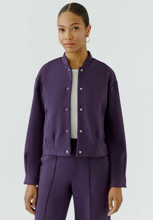 Woman with hoop earrings wearing a buttoned purple jacket and matching pants over a white top, standing against a plain light background.