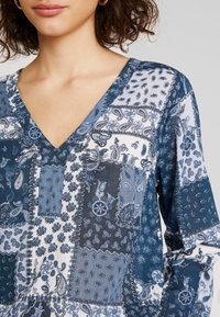 Blouse à motifs bleus avec un décolleté en V. Le tissu présente un mélange de dessins cachemire et floraux. Texture douce avec des nuances de bleu variées.