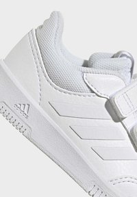 Bílá sportovní obuv s texturovaným koženým svrškem, polstrovaným síťovaným jazykem a suchým zipem. Na boku je zdobena třemi pruhy.