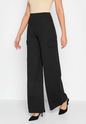 Woman wearing high-waisted black wide-leg pants with side flap pockets and black pointed-toe kitten heels against a white background.