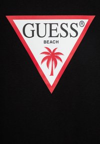 Juodas marškinėliai su baltu ir raudonu trikampiu logotipu. Tekstas sako "GUESS" ir "BEACH", su raudonu palmės grafiką. Lygi tekstūra.