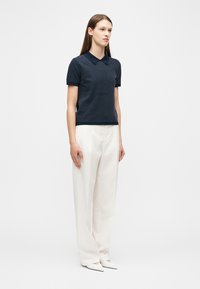 Γυναίκα που στέκεται, φοράει μπλε ναυτικό κοντομάνικο πλεκτό τοπ με λευκές βούλες, λευκό παντελόνι με φαρδιά πόδια και λευκά παπούτσια με μυτερή μύτη.