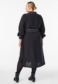 Žena stoji okrenuta leđima, nosi crnu midi haljinu s bijelim pojasom, crne tajice i crne čizme do gležnja, na bijeloj pozadini.