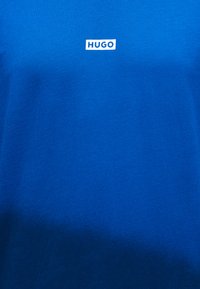 Μπλε ύφασμα με μικρή λευκή ορθογώνια ετικέτα στο κέντρο κοντά στο πάνω μέρος, που δείχνει τη λέξη "HUGO" με έντονα κεφαλαία γράμματα.