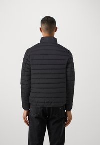 Zwarte pufferjack met horizontaal gestikt ontwerp, hoge kraag en lichte textuur. Draagt over donkere jeans, bekeken vanuit de achterkant.