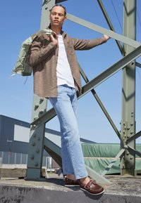 Camicia a quadri marrone sopra una t-shirt bianca, jeans azzurri chiari e scarpe da barca marroni; porta uno zaino verde. Ambiente industriale.