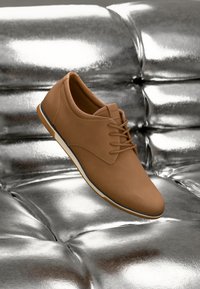 Zapato de cuero sintético marrón con un acabado texturizado, que presenta un diseño de cordones y una suela flexible blanca, colocado sobre un fondo metálico.