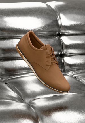 Zapato de cuero sintético marrón con un acabado texturizado, que presenta un diseño de cordones y una suela flexible blanca, colocado sobre un fondo metálico.