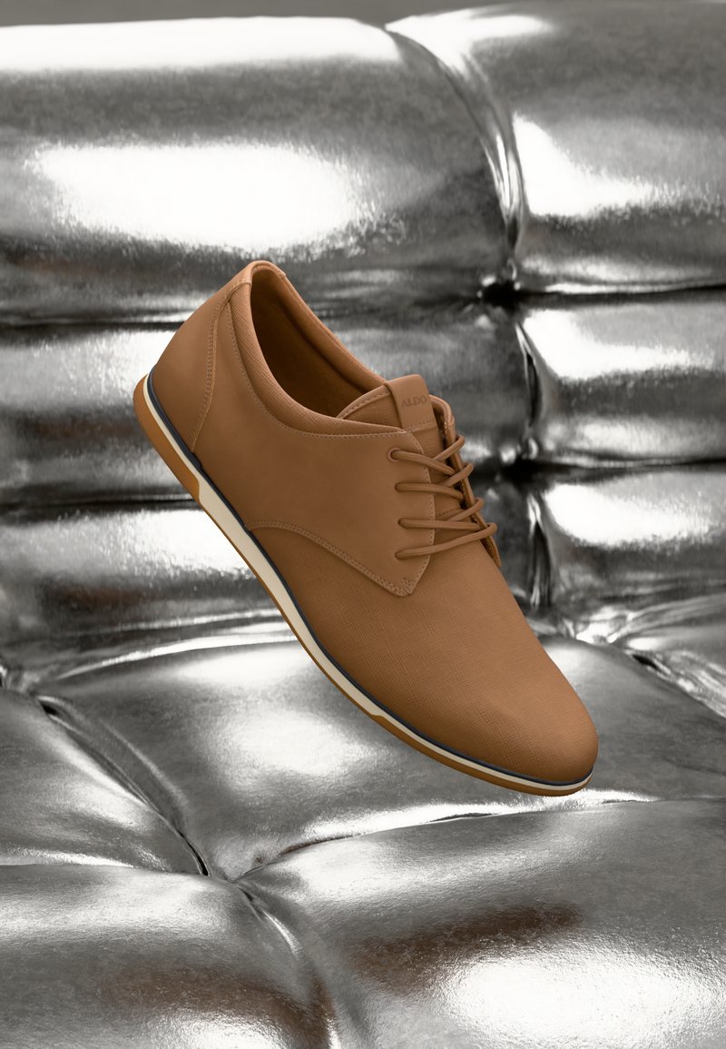 Zapato de cuero sintético marrón con un acabado texturizado, que presenta un diseño de cordones y una suela flexible blanca, colocado sobre un fondo metálico.