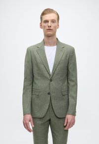 Grüne Leinenjacke mit strukturiertem Finish, zwei Knöpfen, Reverskragen und Seitentaschen, kombiniert mit einem weißen T-Shirt.