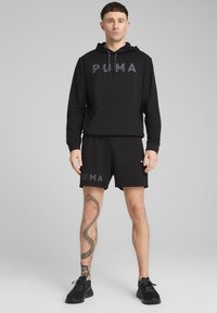 Hanorac negru PUMA cu glugă și buzunar, asortat cu pantaloni scurți negri. Prezintă logo-ul PUMA în gri închis. Modelul poartă adidași negri.
