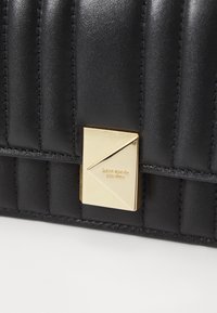 Black quilted leather bag with a gold geometric clasp featuring engraved brand name. Smooth texture and structured design.