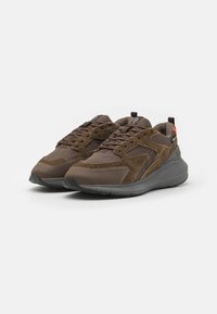 Brown athletic shoes featuring a mix of textured suede and smooth fabric, with a gray sole and orange accent. Lace-up design for fit.