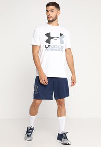 Witte T-shirt met korte mouwen met zwart en grijs Under Armour-logo, gecombineerd met marineblauwe sportshorts met een logoaccent op de zoom.
