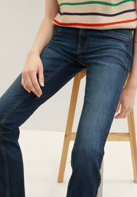 Jean en denim bleu foncé avec une coupe ajustée, présentant des détails de couture subtils et une taille mi-haute, montrés avec une main reposant sur la cuisse.