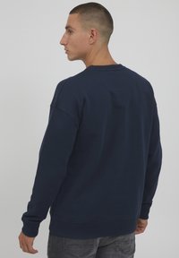 Felpa blu navy con collo rotondo e polsini a costine. Presenta una vestibilità rilassata e una superficie liscia. Vista posteriore che mostra le cuciture.