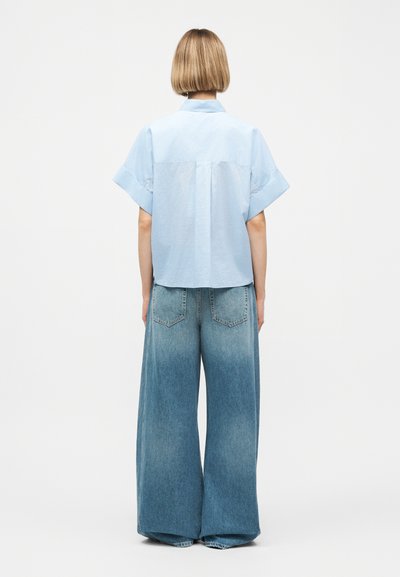 Άτομο με κοντά ξανθά μαλλιά, που φοράει ανοιχτό μπλε κοντομάνικο πουκάμισο και φαρδιά ξεβαμμένα μπλε τζιν, στέκεται αντικρίζοντας ένα απλό λευκό φόντο.