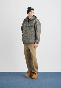 Bunda s maskáčovým vzorom, ľahký materiál, kapucňa so šnúrkou, predná vrecko; kombinovaná s béžovými nohavicami a žltými pracovnými čižmami.