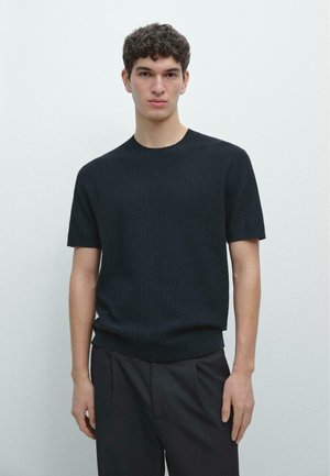 Giovane uomo con capelli corti e ricci che indossa un maglione nero a maniche corte e pantaloni neri, in piedi davanti a uno sfondo chiaro e uniforme.