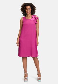Magenta ärmelloses Kleid mit einer Schleifendetail an der Schulter, aus weichem Stoff gefertigt. Das Model trägt silberne Plateausandalen mit glänzender Oberfläche.