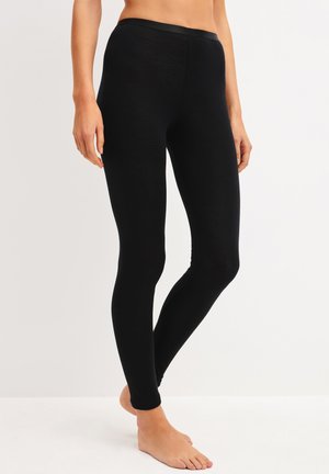 Persoon draagt effen zwarte legging met volle lengte en staat blootsvoets tegen een effen witte achtergrond.