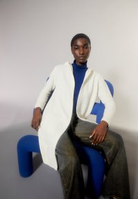 Weißer langer Cardigan über einem blauen Rollkragenpullover, kombiniert mit lockeren dunkelblauen Jeans. Das Model sitzt auf einem blauen strukturierten Stuhl vor einem neutralen Hintergrund.