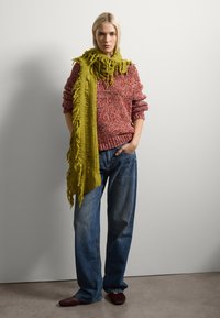 Suéter rojo y multicolor de punto con mangas enrolladas, combinado con jeans de pierna ancha de color azul. Acentuado por una bufanda verde larga y deshilachada y zapatos de color burdeos.