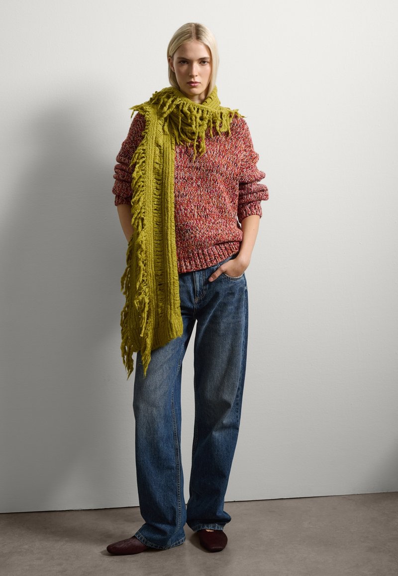 Suéter rojo y multicolor de punto con mangas enrolladas, combinado con jeans de pierna ancha de color azul. Acentuado por una bufanda verde larga y deshilachada y zapatos de color burdeos.