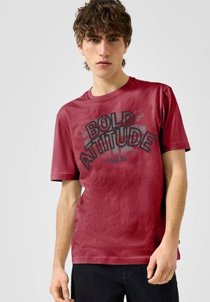 Junger Mann mit lockigem Haar, der ein rotes T-Shirt mit der Aufschrift "BOLD ATTITUDE" in großen schwarzen Buchstaben trägt, vor einem schlichten Hintergrund steht.