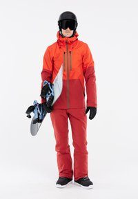 Männliches rotes Snowboard-Outfit mit einer wasserdichten Jacke mit Reißverschlüssen, passenden Hosen, Handschuhen und einem Snowboardhelm, der ein Snowboard hält.