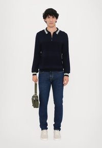Tumedane kootud pullover, millel on lukuga kaelus, roheliste ja valgete triipudega, koos siniste teksade ja beežide tossudega, hoides oliivroheline kotti.