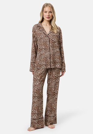 Kobieta stoi boso, ubrana w koszulę z długim rękawem zapinaną na guziki oraz szerokie spodnie w brązowo-czarne cętki leopardzie.