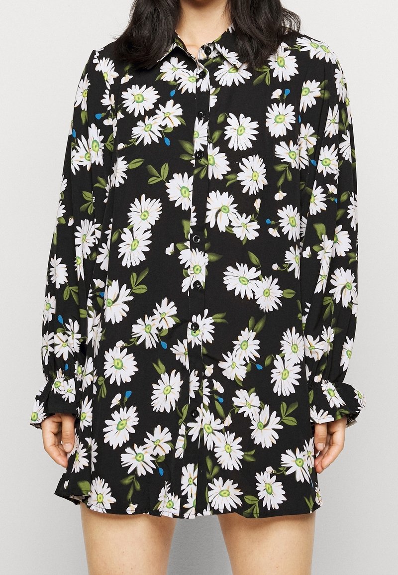 Robe chemise noire à manches longues avec imprimé marguerites blanches, feuilles vertes et petits accents bleus, portée par une personne visible du cou à mi-cuisse.