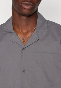 Šedá krátká košile s uvolněným límcem, jednou náprsní kapsou a knoflíkovým zapínáním. Hladká tkanina s jemnou strukturou. Viditelný stříbrný náhrdelník.