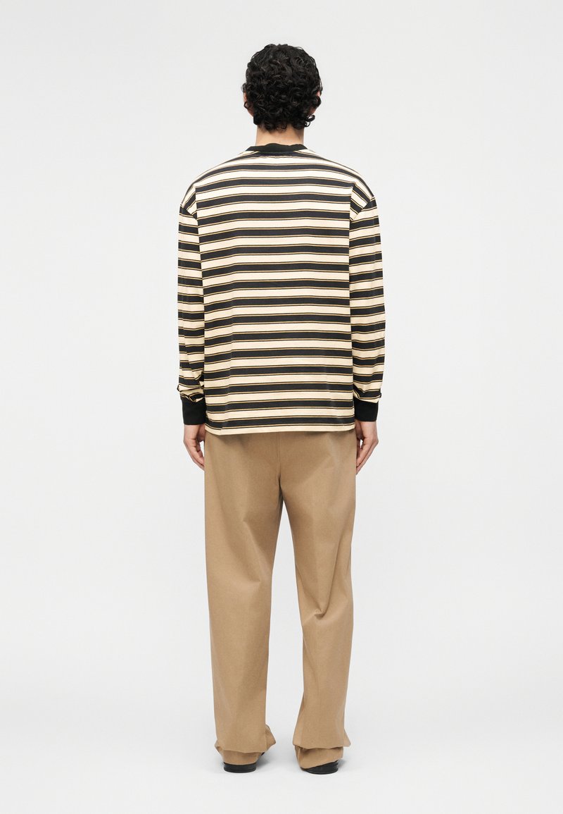 Man met krullend haar, gekleed in een zwart-beige gestreept shirt met lange mouwen en loszittende beige broek, staand met de rug naar voren.