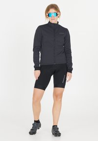 Svart cykeljacka med hög krage, långa ärmar och dragkedja. Kombinationen med svarta shorts och cykelskor, har reflektorer.