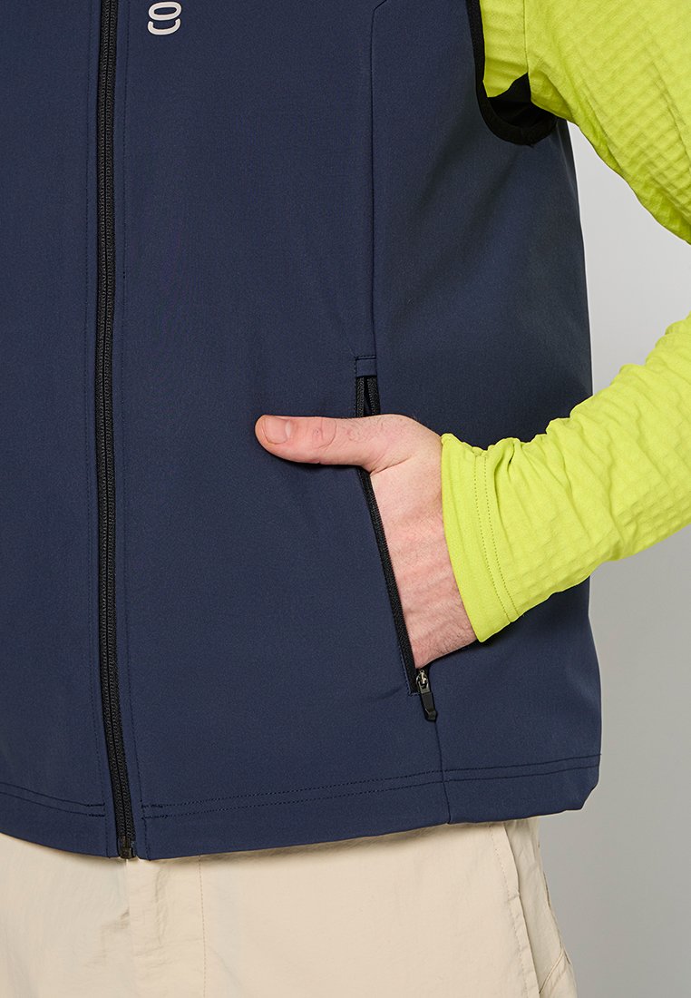 Gilet blu navy con chiusura a zip e tasca laterale, indossato su una camicia a maniche lunghe verde brillante. Tessuto liscio, taglio atletico, minimalismo nel marchio.